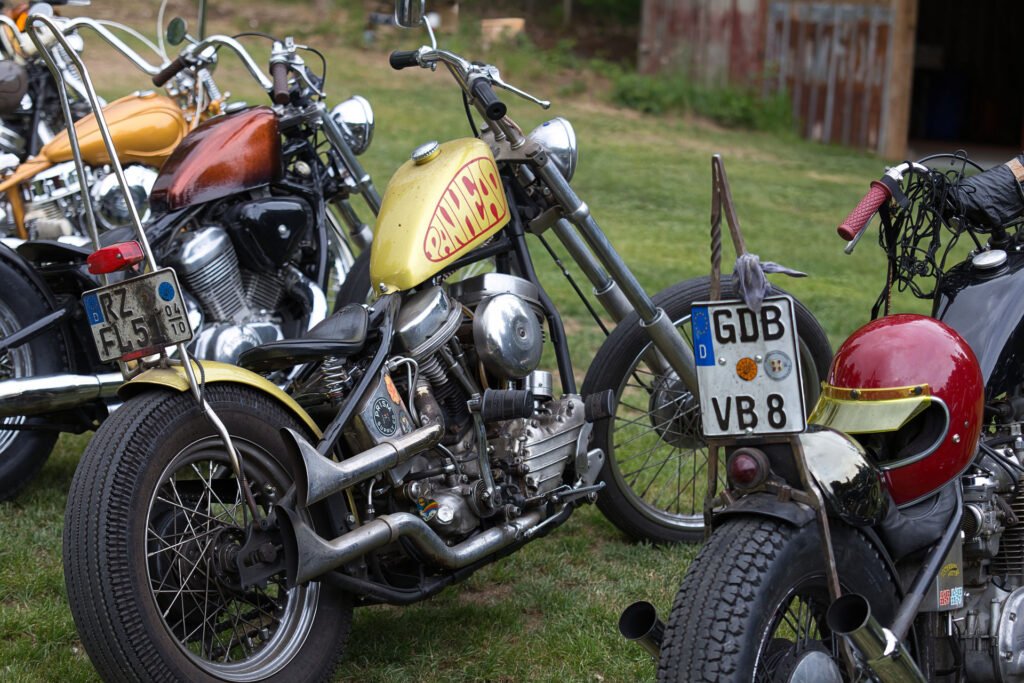 Drei Oldtimer-Motorräder mit Sonderlackierung und Nummernschildern sind auf einer Wiese geparkt. Das mittlere Motorrad ist gelb mit dem Wort "Sanchez" auf dem Tank. Im Hintergrund ist ein rustikales Holzgebäude zu sehen.