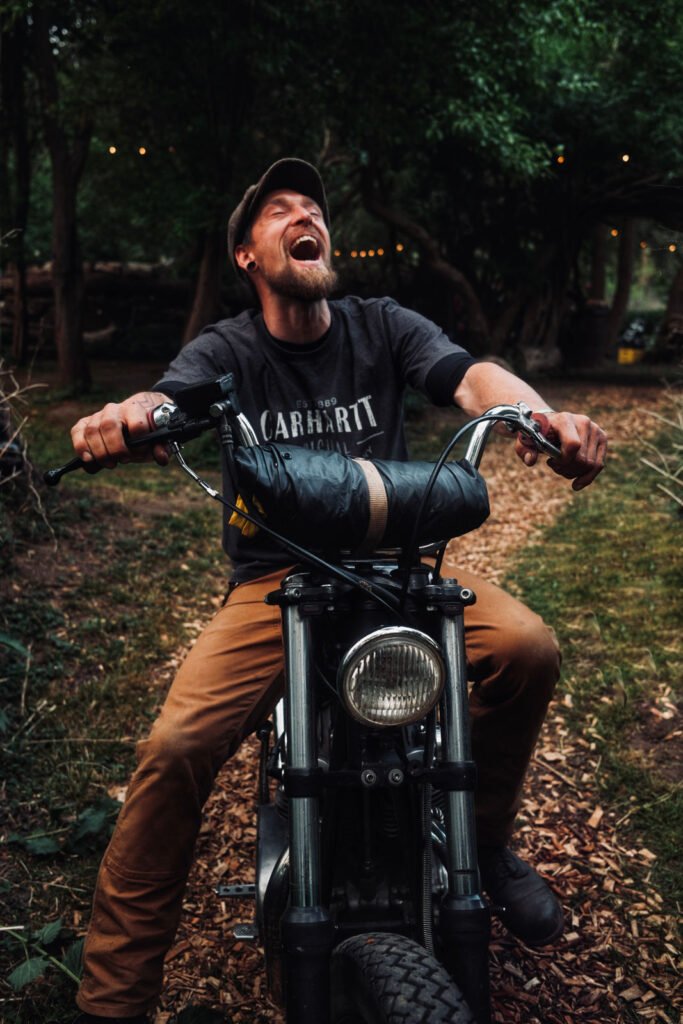 Ein Mann mit Bart und Mütze lacht fröhlich, während er auf einem geparkten Motorrad in einem bewaldeten Außenbereich sitzt. Er trägt ein dunkles Carhartt-Hemd und eine braune Hose.