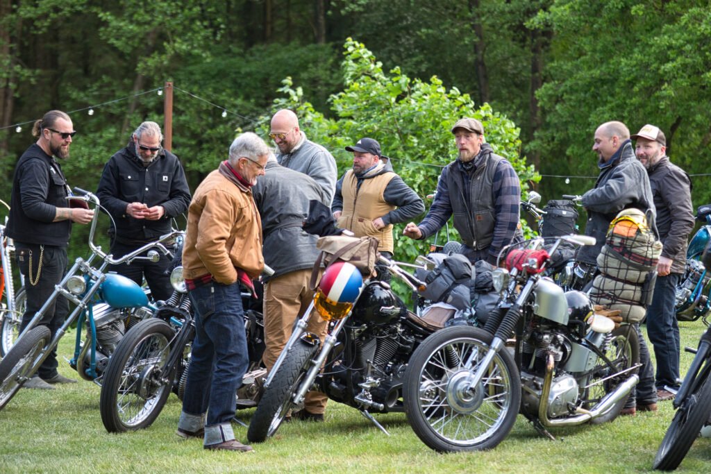 Eine Gruppe von Männern steht im Freien neben mehreren geparkten Motorrädern und unterhält sich auf einer von Bäumen umgebenen Wiese. Einige tragen Jacken und Hüte; an den Motorrädern sind Taschen festgeschnallt.