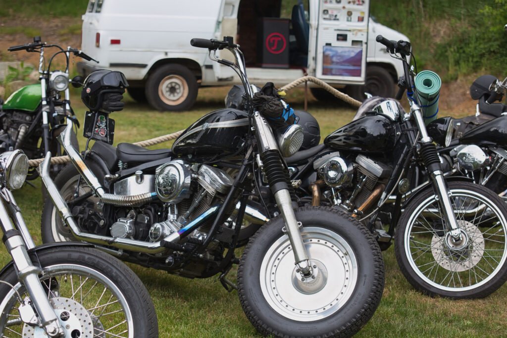 Mehrere geparkte Motorräder, meist schwarz mit verchromten Details, stehen auf einer Wiese in der Nähe eines weißen Lieferwagens und von Außenaufstellern. Auf einem der Motorräder befindet sich ein Helm, an einem anderen ist eine grüne Matte befestigt.