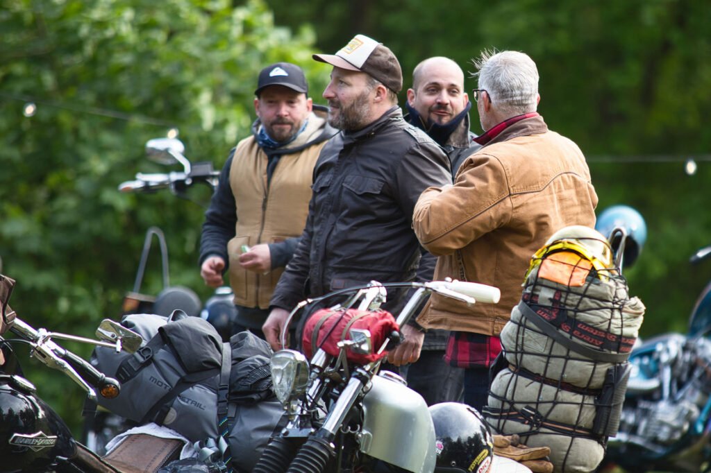 Vier Männer stehen im Freien zwischen Motorrädern, die mit Reiseutensilien beladen sind, tragen Jacken und Hüte, unterhalten sich und lächeln, während im Hintergrund grüne Bäume zu sehen sind.