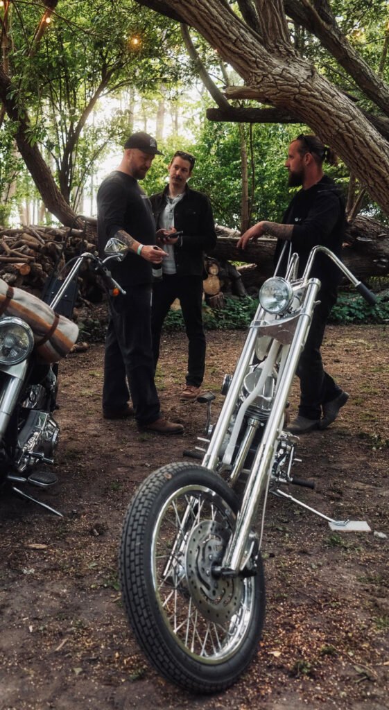 Drei Männer stehen im Freien neben Bäumen und Motorrädern und unterhalten sich. Ein Motorrad ist im Vordergrund zu sehen, während gestapeltes Brennholz und Grünpflanzen den Hintergrund füllen.