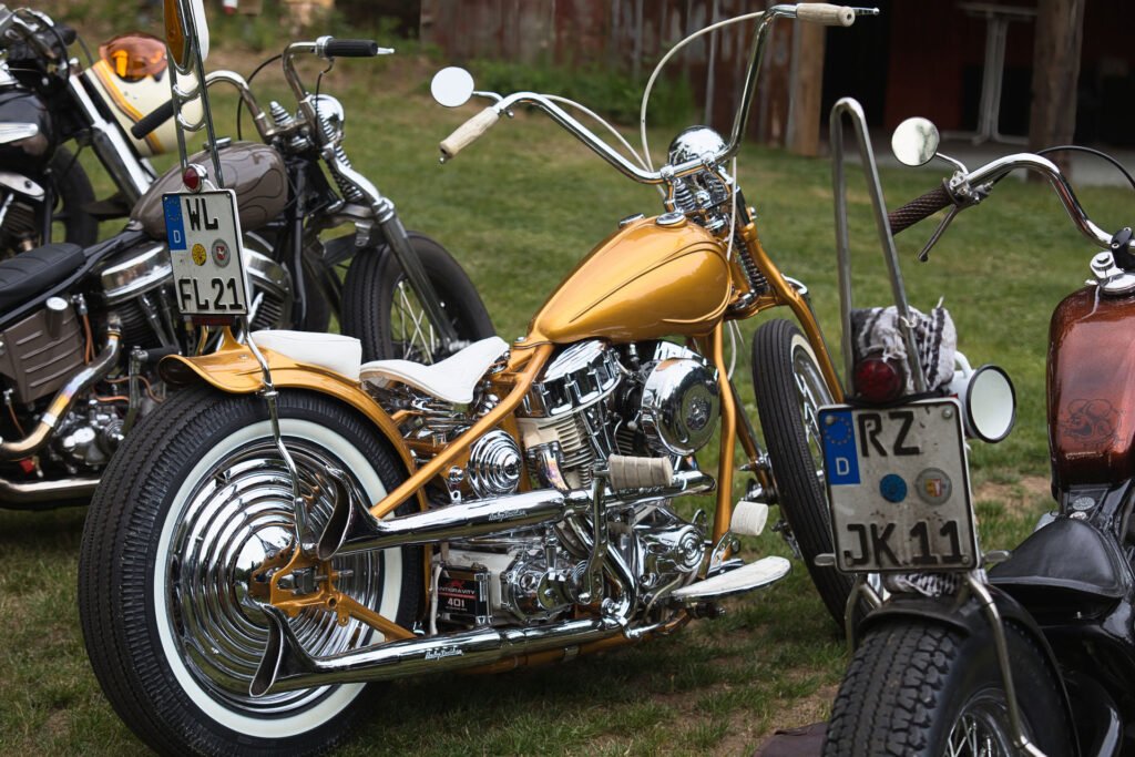 Ein goldglänzendes Custom-Chopper-Motorrad mit verchromten Details und weißer Sitzbank ist bei einer Outdoor-Veranstaltung zwischen anderen Motorrädern auf dem Rasen geparkt. Das Motorrad hat einen hohen Lenker und ein deutsches Nummernschild.