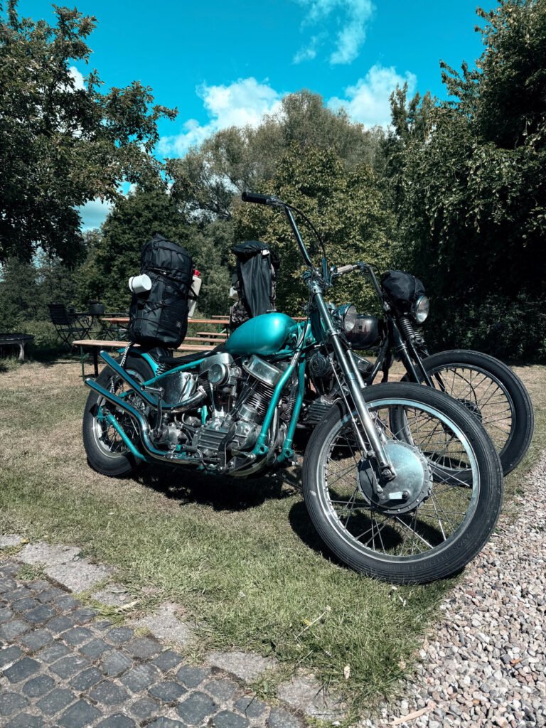 Zwei Oldtimer-Motorräder mit hohen Lenkern sind auf einer Wiese in der Nähe eines Kopfsteinpflasters geparkt, auf deren Sitzen Rucksäcke befestigt sind. An einem sonnigen Tag sind im Hintergrund Bäume und ein Picknicktisch zu sehen.