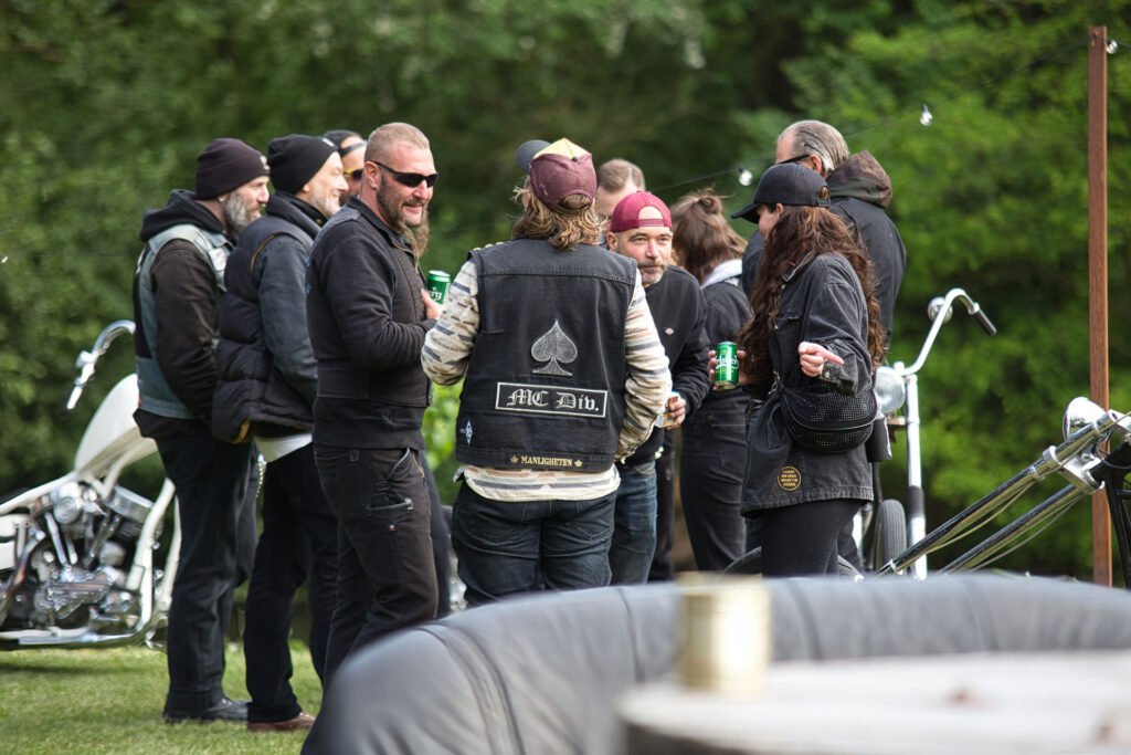Eine Gruppe von Bikern mit Jacken und Mützen steht im Freien in einem Kreis, unterhält sich und hält Getränke in der Hand. Zwei weiße Motorräder sind in der Nähe geparkt, und im Hintergrund sind Bäume zu sehen.