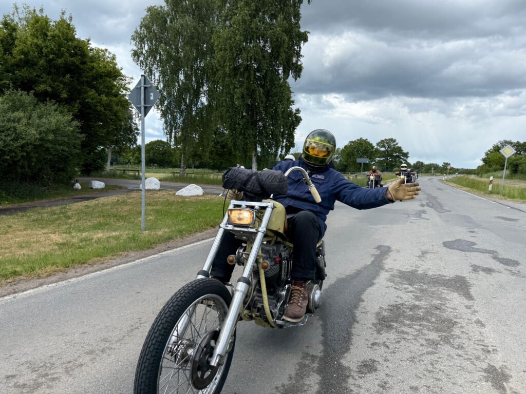 Eine Person mit Helm und Handschuhen fährt auf einem Motorrad eine Landstraße entlang und streckt einen Arm in einer freundlichen Geste zur Seite aus. Bäume und Gras säumen die Straße unter einem bewölkten Himmel. Andere Motorradfahrer folgen ihm.