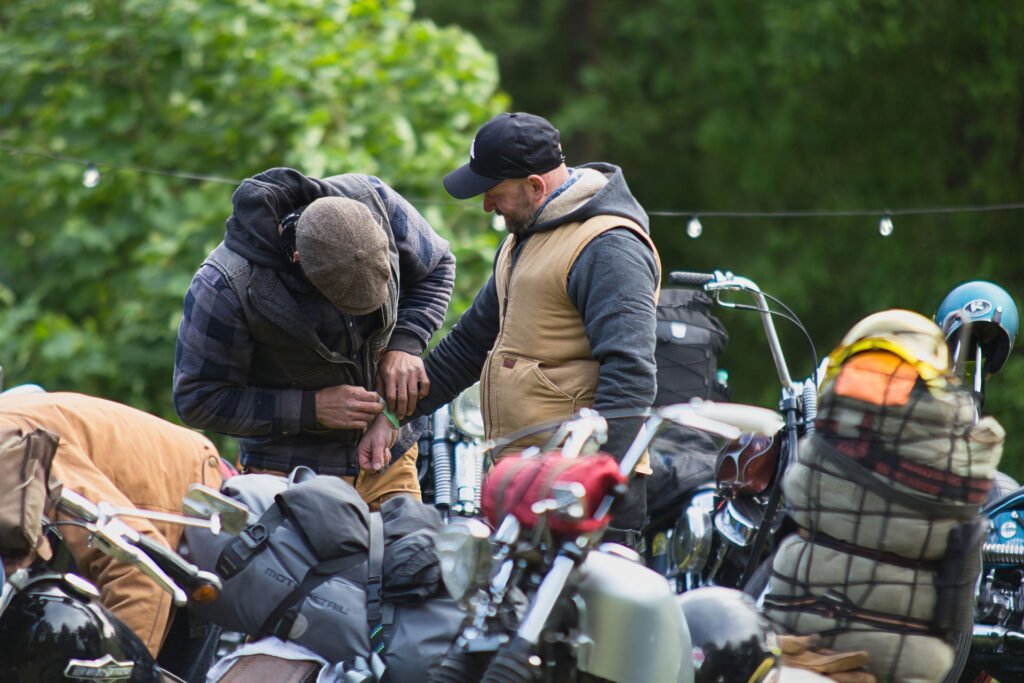 Zwei in Outdoor-Kleidung gekleidete Personen stehen neben Motorrädern, die mit Ausrüstung beladen sind. Eine Person justiert etwas an ihrer Jacke, während die andere zusieht, umgeben von Grünzeug und Lichterketten im Hintergrund.