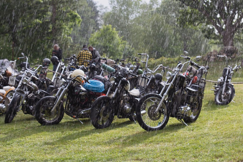 Eine Gruppe von Motorrädern ist im Regen auf einer Wiese geparkt, und mehrere Personen stehen zwischen den Motorrädern. Im Hintergrund sind Bäume und Grünflächen zu sehen.