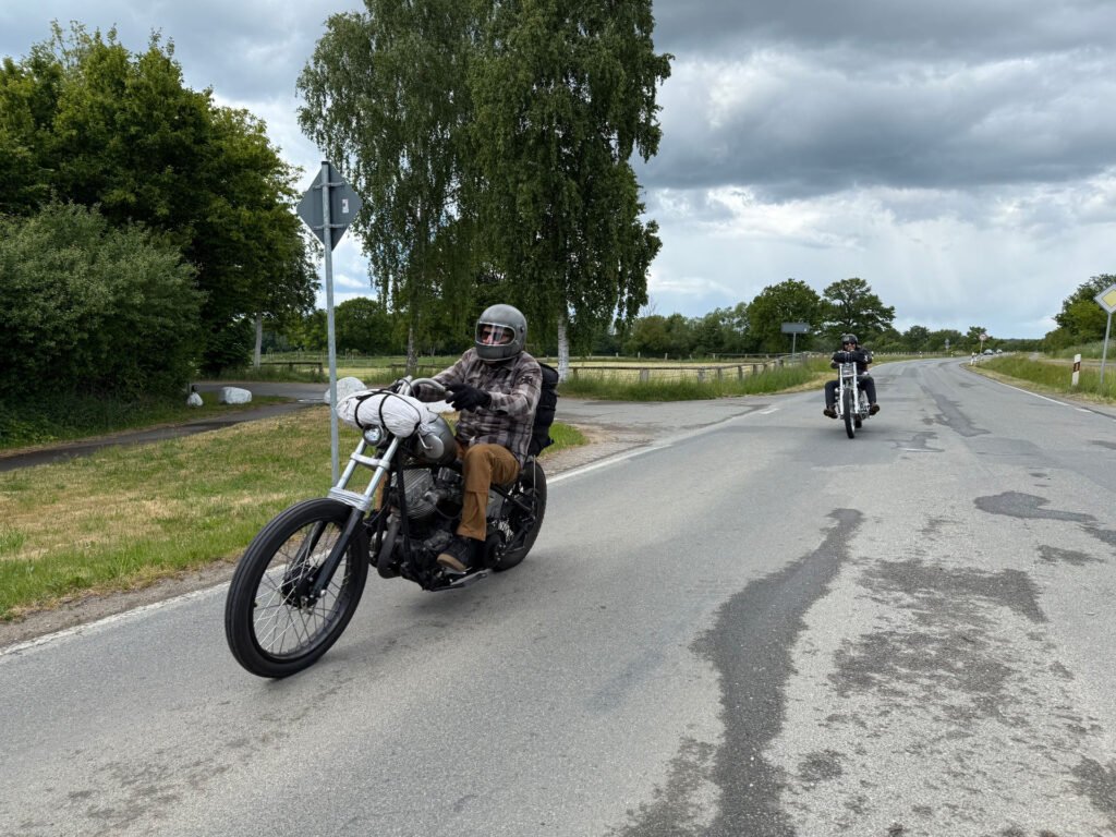 Zwei Personen fahren mit ihren Motorrädern auf einer Landstraße, die von grünen Bäumen und Gras gesäumt ist, unter einem bewölkten Himmel. Der Fahrer vor ihnen trägt ein kariertes Hemd, einen Helm und eine Sonnenbrille. Die Straße macht eine Kurve und ist auf beiden Seiten begrünt.
