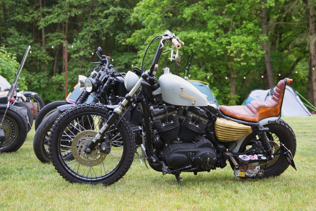 Ein Custom-Motorrad mit Geländereifen, einem braunen Ledersitz und einem hellgrauen Kraftstofftank ist im Gras geparkt. Andere Motorräder und Zelte sind im Hintergrund zwischen grünen Bäumen zu sehen.
