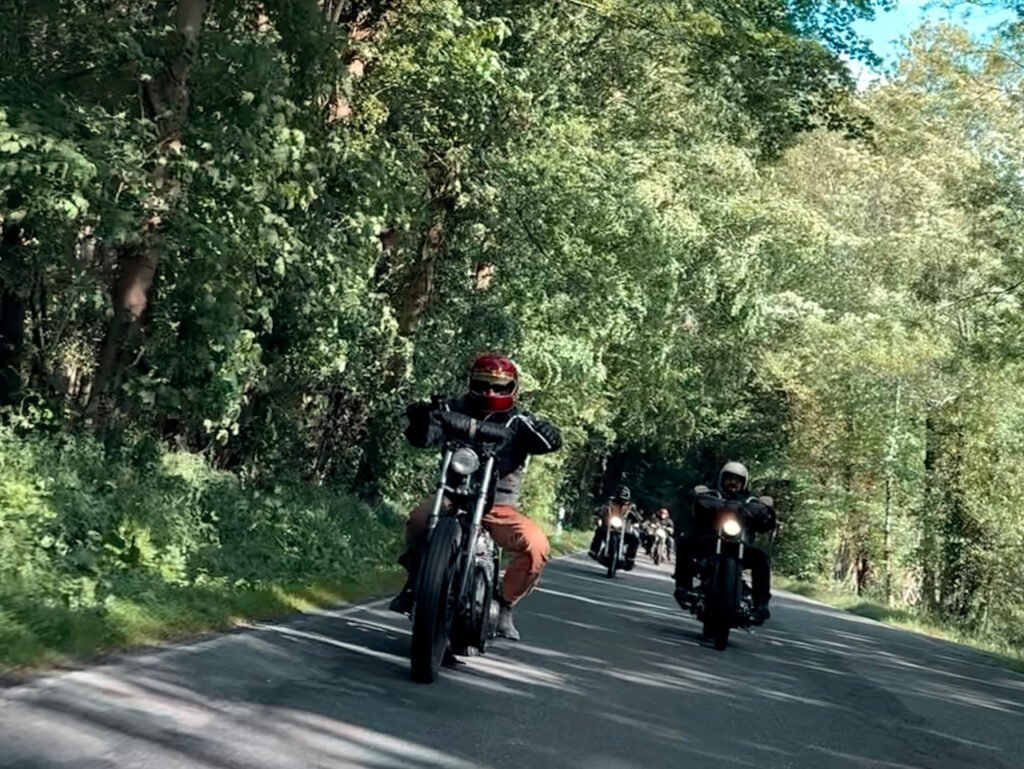 Eine Gruppe von Motorradfahrern fährt an einem sonnigen Tag eine von Bäumen gesäumte Straße hinunter, wobei der führende Fahrer einen roten Helm und braune Hosen trägt. Üppiges grünes Laub umgibt die Straße.