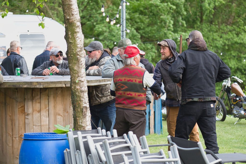Eine Gruppe von Männern versammelt sich im Freien an einem hölzernen Bartresen, unterhält sich und schüttelt Hände. Einige tragen Hüte und Jacken. Im Hintergrund sind Bäume, ein blaues Fass, Stuhlreihen und ein Wohnwagen zu sehen.