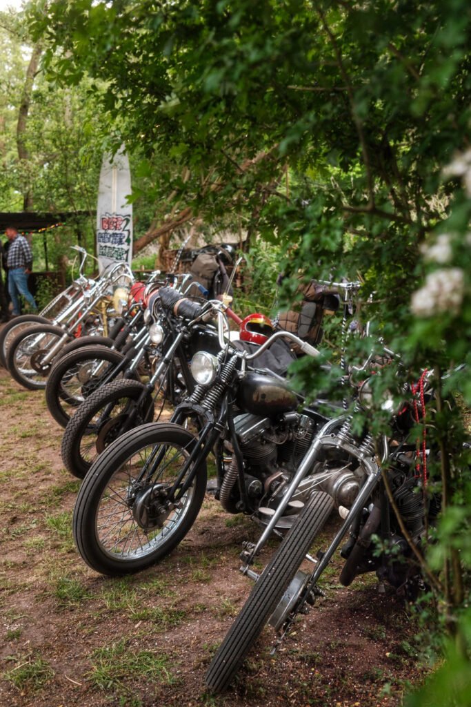 Eine Reihe von Oldtimer-Motorrädern, die auf einem unbefestigten Weg in einem Waldgebiet geparkt sind, umgeben von Bäumen und Grünpflanzen. Eine Person steht im Hintergrund in der Nähe eines Banners.