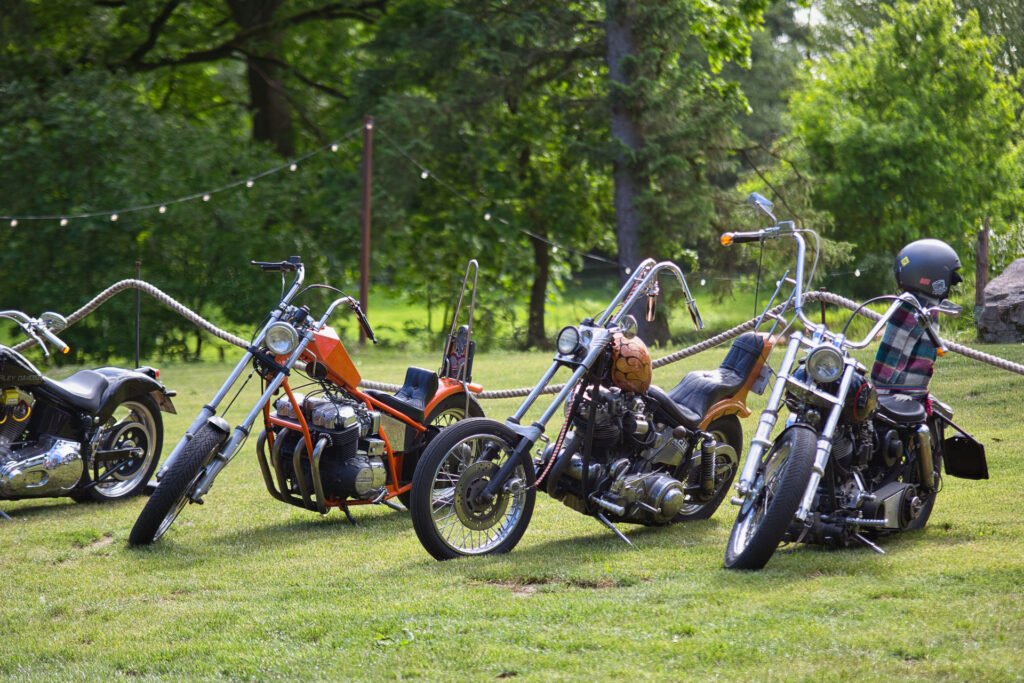 Mehrere Custom-Motorräder sind auf einer Wiese in einem bewaldeten Gebiet geparkt, auf einem Motorrad sitzt eine Person mit Helm und kariertem Hemd. Im Hintergrund sind Lichterketten und Bäume zu sehen.