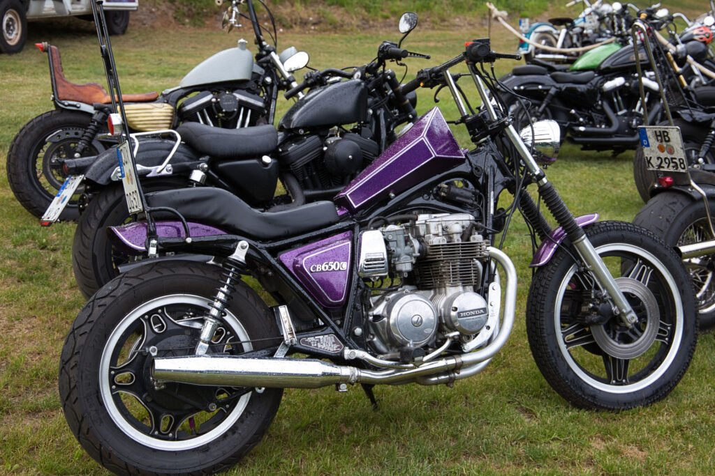Eine Gruppe geparkter Motorräder auf einer Wiese, mit einer lila Honda CB650 Custom im Vordergrund und anderen Motorrädern im Hintergrund.