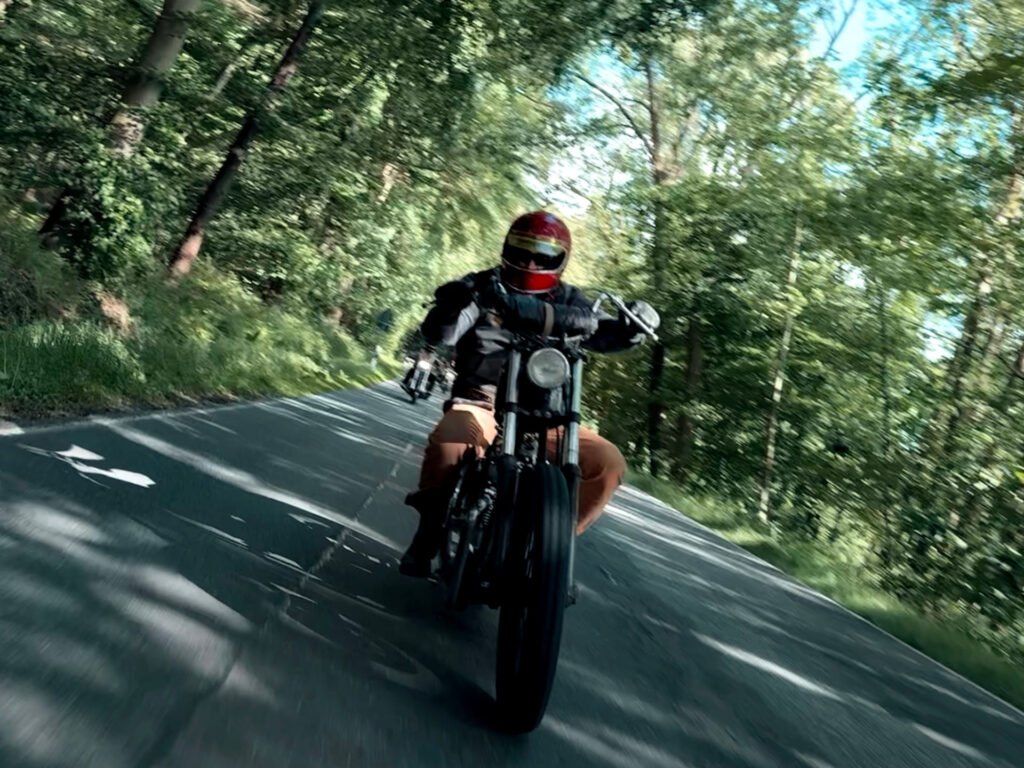 Eine Person mit rotem Helm und schwarzer Jacke fährt an einem sonnigen Tag auf einem Motorrad eine von Bäumen gesäumte Straße hinunter und neigt sich beim Durchfahren der Kurve leicht nach links.