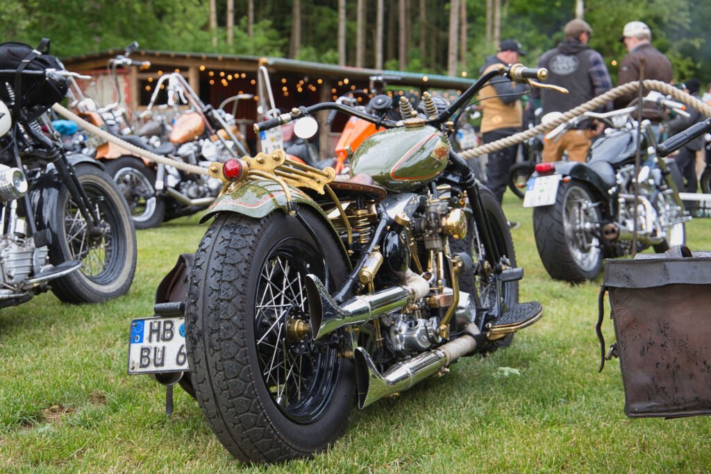 Ein Oldtimer-Motorrad mit verchromten und goldenen Details ist auf einer Wiese zwischen anderen Motorrädern geparkt. Menschen stehen in der Nähe, und im Hintergrund steht ein Holzpavillon mit Lichterketten. Die Szene ist von Bäumen umgeben.