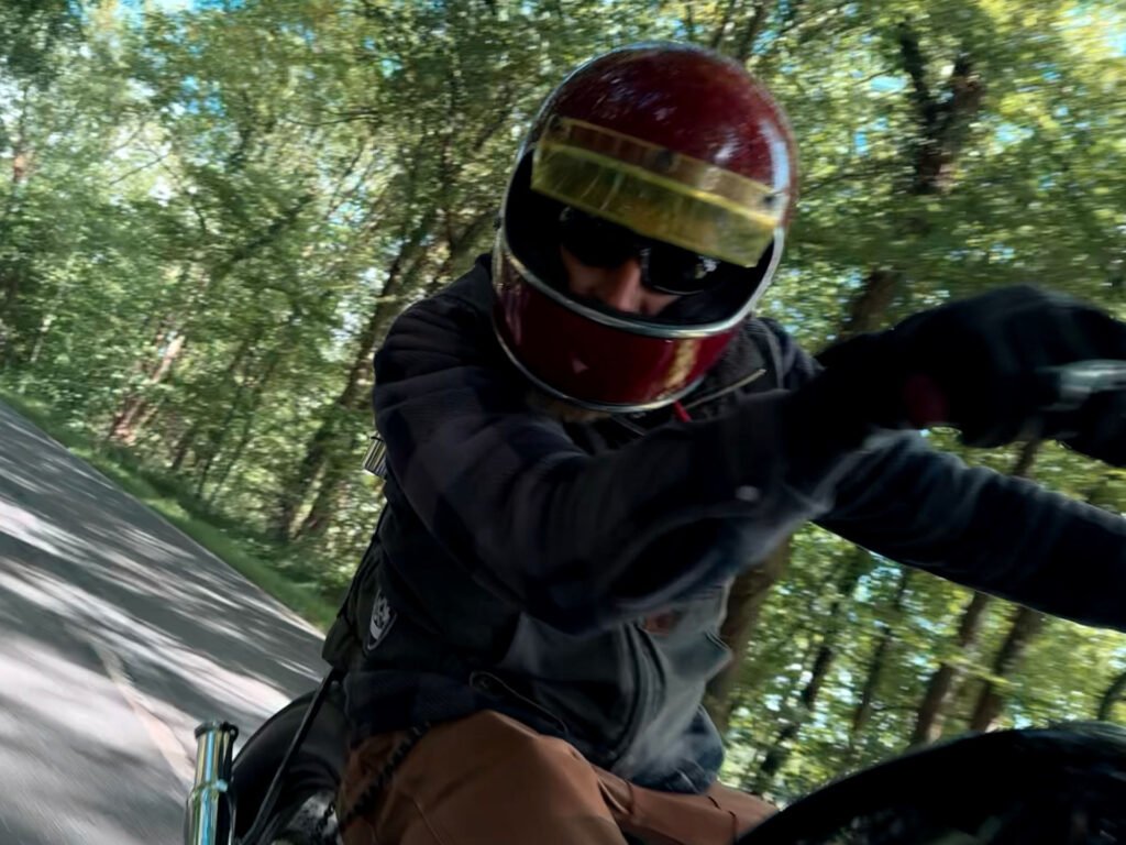 Eine Person, die einen roten Helm und eine Sonnenbrille trägt, fährt auf einem Motorrad schnell eine von Bäumen gesäumte Straße hinunter und biegt in eine Kurve ein. Das Sonnenlicht fällt durch das grüne Laub über ihr.