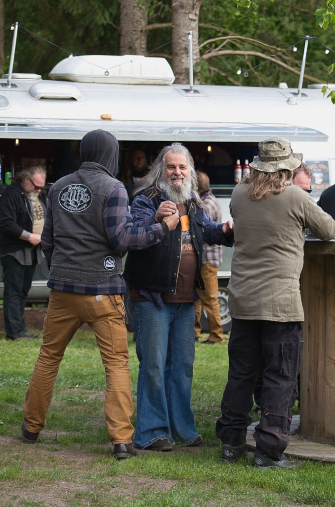 Eine Gruppe von Männern steht im Freien neben einer hölzernen Bar und einem silbernen Imbisswagen. Ein Mann mit langen grauen Haaren und einem Bart, der eine Jeansweste und Schlagjeans trägt, lächelt, während er einem anderen Mann in einer Kapuzenjacke die Hand schüttelt.