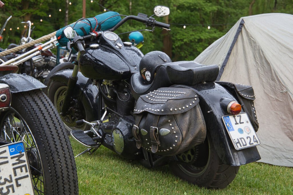 Ein schwarzes Motorrad mit beschlagenen Ledertaschen ist auf einer Wiese in der Nähe eines Zeltes geparkt, auf dessen Oberfläche Regentropfen fallen. Im Hintergrund sind weitere Motorräder und Lichterketten zu sehen.