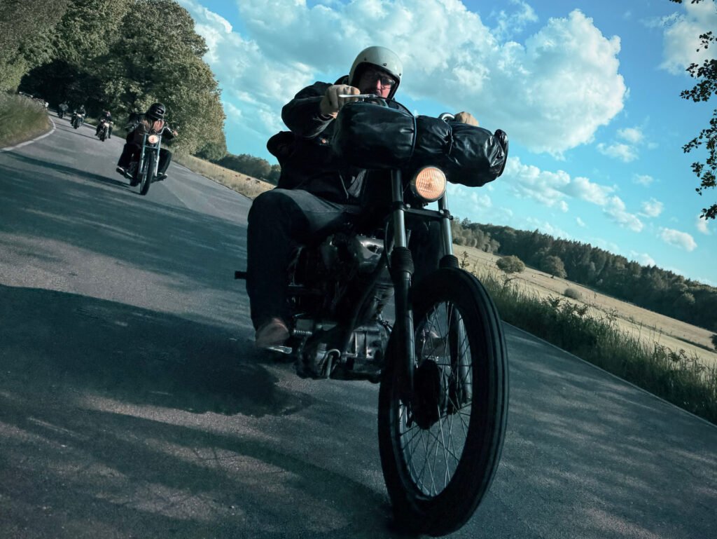 Eine Person, die einen Helm trägt, fährt mit einem Oldtimer-Motorrad auf einer kurvenreichen Landstraße, gefolgt von anderen Motorradfahrern. Bäume säumen eine Seite der Straße unter einem teilweise bewölkten blauen Himmel.