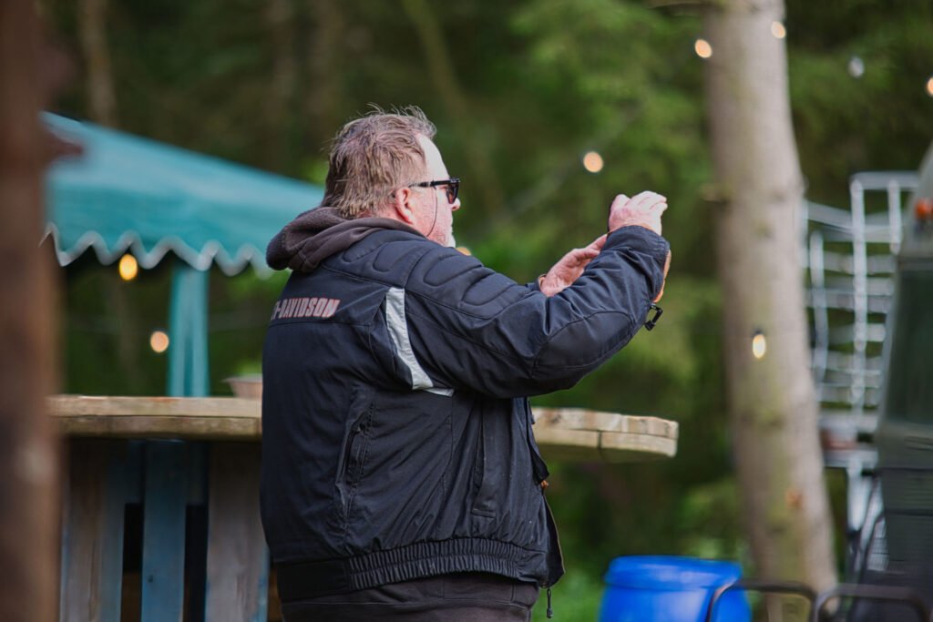Ein Mann in einer schwarzen Harley-Davidson-Jacke steht im Freien an einem Holztresen und hält etwas in den Händen. Im Hintergrund sind Bäume und Lichterketten zu sehen.