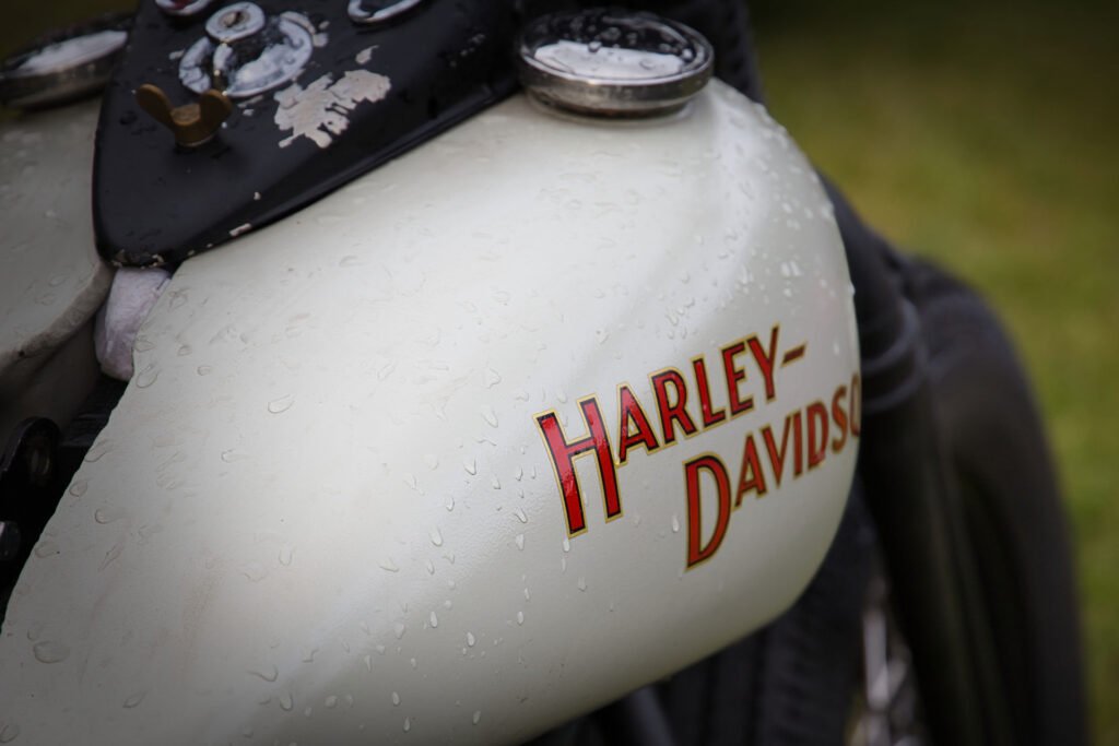Nahaufnahme eines alten Harley-Davidson Motorradtanks mit Wassertropfen auf seiner weißen Oberfläche und dem Markennamen in roten und goldenen Buchstaben.