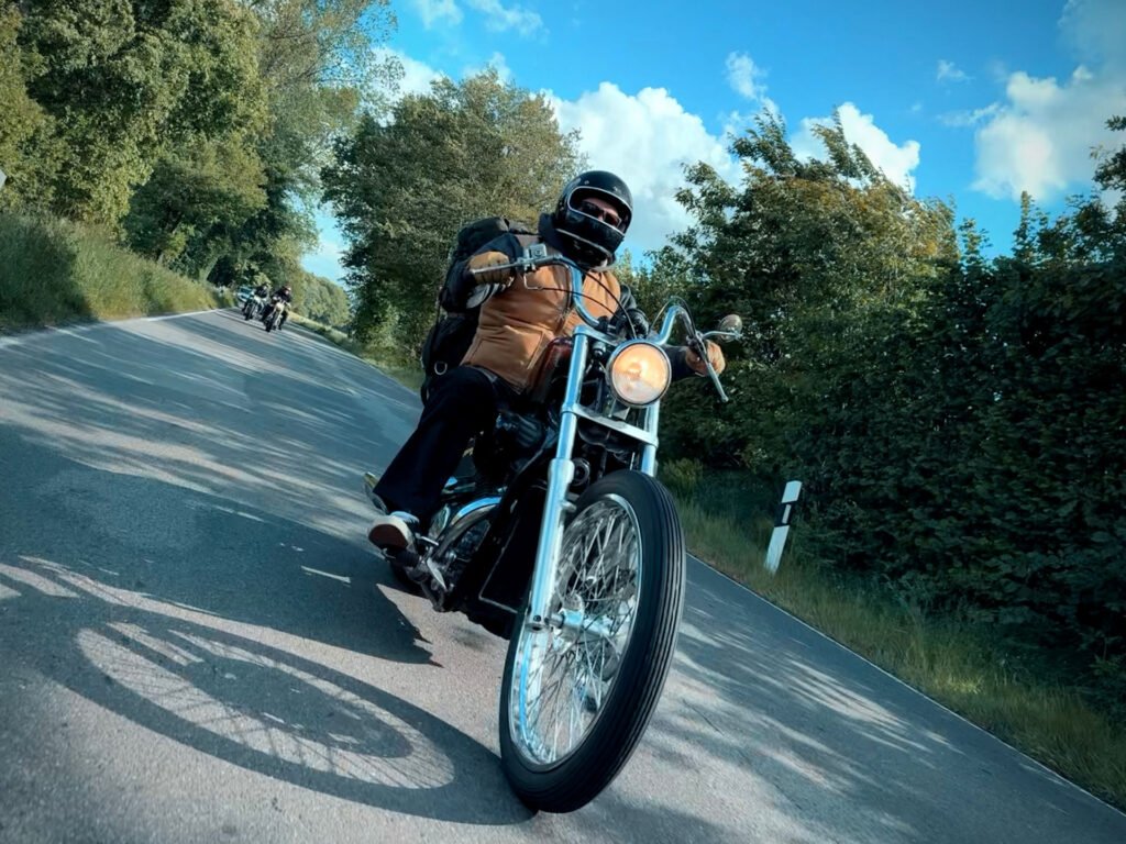 Eine Person, die einen Helm und einen Rucksack trägt, fährt auf einer von Bäumen gesäumten Landstraße mit anderen Motorradfahrern, die in der Ferne unter einem blauen Himmel mit Wolken zu sehen sind.