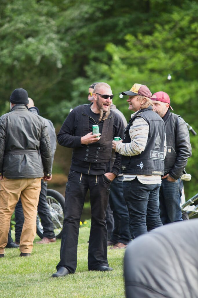 Zwei Männer, beide mit Motorradwesten und Getränken in der Hand, stehen im Gras und lachen zusammen bei einem Treffen im Freien. Im Hintergrund mit grünen Bäumen sind weitere Personen in Jacken und ein Motorrad zu sehen.