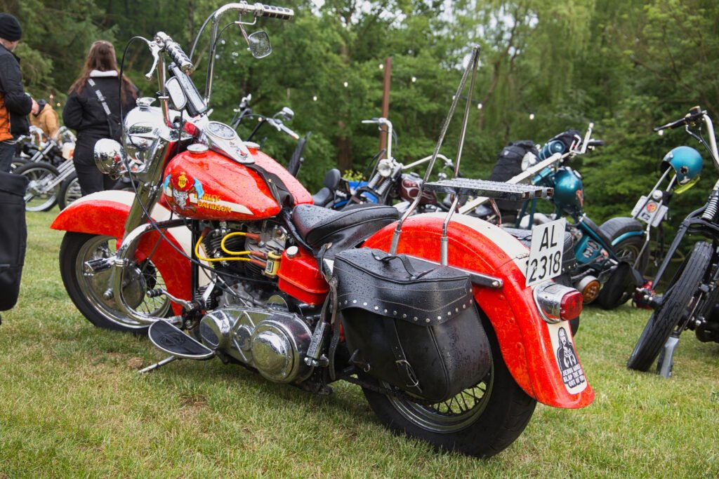 Ein leuchtend rotes Oldtimer-Motorrad mit Ledersatteltaschen ist auf einer Wiese zwischen anderen Motorrädern geparkt. Das Motorrad ist nass vom Regen, und im Hintergrund sind Bäume zu sehen.