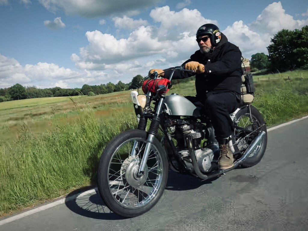 Ein Mann mit Helm und Sonnenbrille fährt auf einem Oldtimer-Motorrad eine Landstraße entlang, mit grünen Feldern und Bäumen im Hintergrund unter einem teilweise bewölkten Himmel.