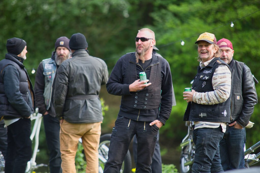 Eine Gruppe von Männern mit Jacken und Hüten steht im Freien, unterhält sich und lacht. Einige halten Dosen, möglicherweise Getränke, und im Hintergrund sind teilweise einige Motorräder zu sehen. Sie sind von Bäumen und Grünflächen umgeben.