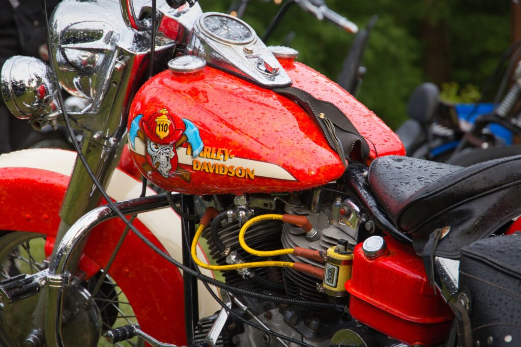 Nahaufnahme eines roten Harley-Davidson Motorrads mit Regentropfen auf dem Tank, mit einer bunten Adlergrafik und dem Harley-Davidson Logo; Teile des Motors und des Lenkers sind sichtbar.