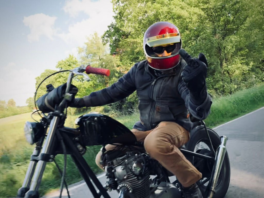 Ein Motorradfahrer mit rotem Helm, schwarzen Handschuhen und braunen Hosen fährt auf einer Landstraße, streckt den linken Arm aus und hebt den Mittelfinger in Richtung der Kamera. Grüne Bäume und Gras säumen den Straßenrand.