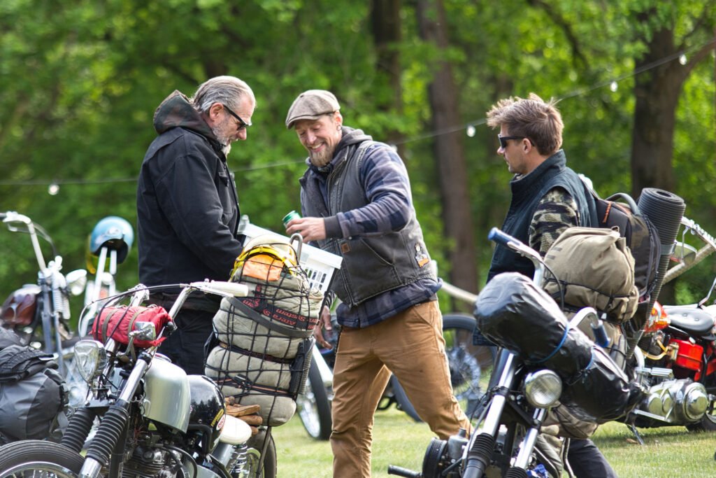 Drei Männer stehen neben ihren geparkten Motorrädern im Freien, unterhalten sich und lächeln. Die Motorräder sind mit Reiseutensilien beladen, und im Hintergrund sind Bäume mit grünem Laub zu sehen. Es scheint ein zwangloses, freundliches Treffen zu sein.