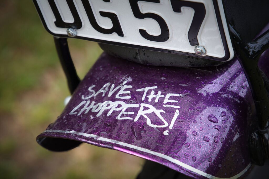 Nahaufnahme eines lilafarbenen Motorradkotflügels mit Regentropfen und der handschriftlichen Botschaft "SAVE THE CHOPPERS!" unter einem teilweise sichtbaren Nummernschild.