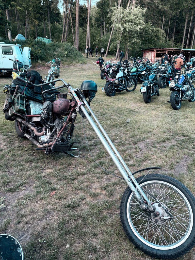 Ein langgestrecktes Custom-Chopper-Motorrad ist auf einer Wiese bei einem Treffen mit mehreren anderen Motorrädern und Menschen im Hintergrund geparkt, umgeben von Bäumen und Wald.
