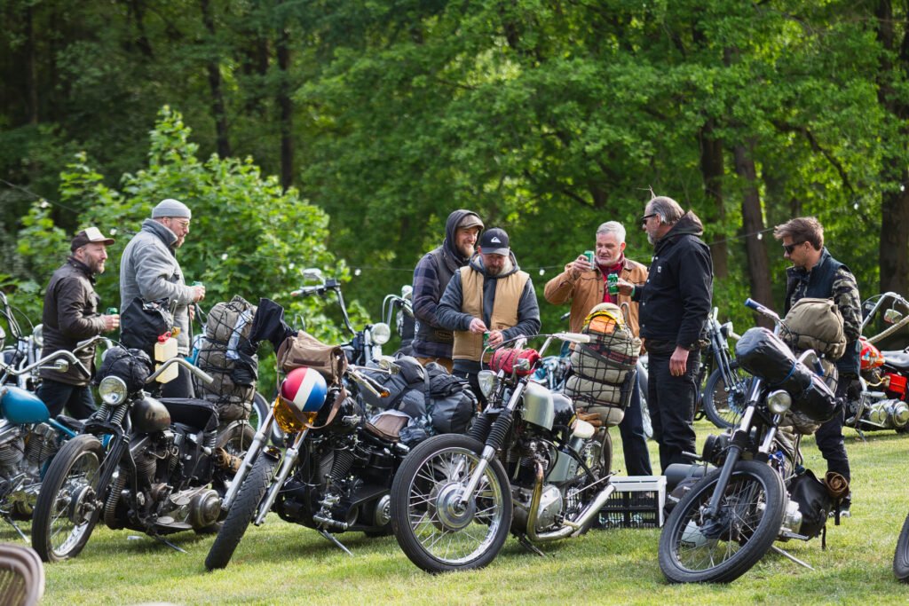 Eine Gruppe von Menschen steht im Freien neben mehreren geparkten Motorrädern, unterhält sich und lächelt. Sie sind mit Jacken und Hüten bekleidet, umgeben von Gras und Bäumen, was auf ein zwangloses Treffen oder ein Motorradtreffen schließen lässt.