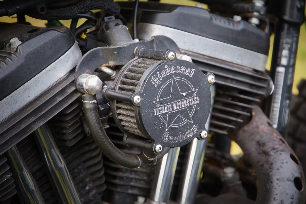 Nahaufnahme eines Motorradmotors mit einer speziell angefertigten Luftfilterabdeckung mit der Aufschrift "Outcast Rebel Riders", verziert mit einem Stern und einem Schraubenschlüssel, mit Schrauben und Metallrohren, mit Anzeichen von Rost und Verschleiß.