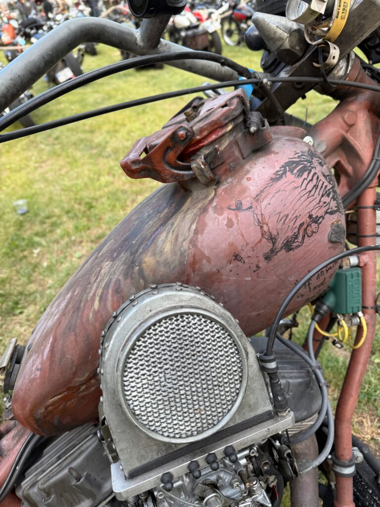Eine Nahaufnahme eines verwitterten, rostigen Motorradtanks mit verblasster Farbe, sichtbaren Dellen und kundenspezifischen Metallteilen, geparkt auf einer Wiese mit anderen Motorrädern im unscharfen Hintergrund.