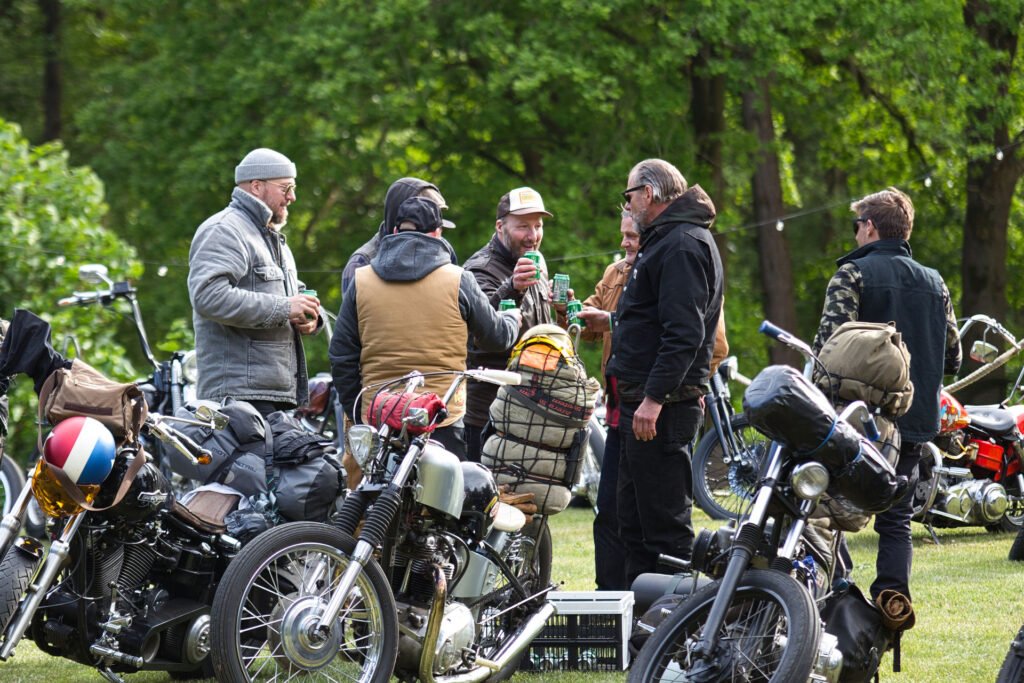 Eine Gruppe von Männern mit Jacken und Hüten steht im Freien neben mehreren geparkten Motorrädern, unterhält sich und hält Getränke in der Hand, während im Hintergrund grüne Bäume stehen.