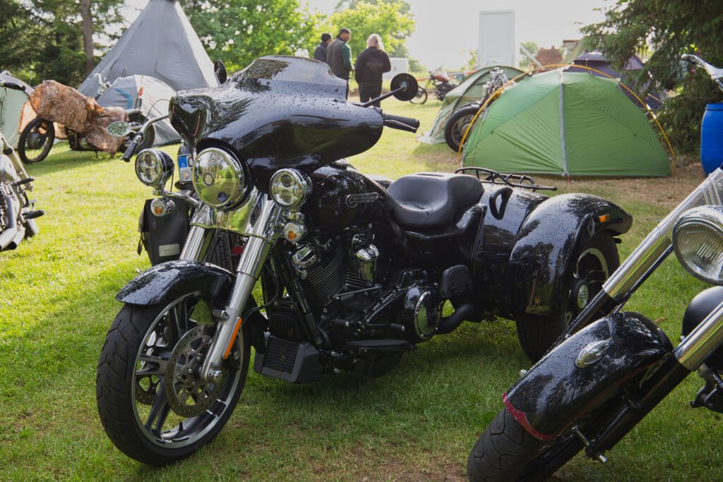 Ein schwarzes, glänzendes, dreirädriges Motorrad ist auf einer Wiese in der Nähe von Zelten geparkt, im Hintergrund stehen ein paar Leute und unterhalten sich. Das Motorrad ist nass, was auf kürzlichen Regen hindeutet. Auch andere Motorräder sind in der Nähe zu sehen.