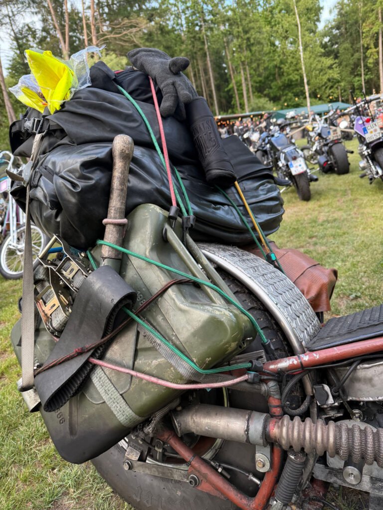 Nahaufnahme eines Motorrads, das mit einer Campingausrüstung beladen ist, darunter ein Kanister, eine Axt, schwarze Taschen, Handschuhe und elastische Schnüre, geparkt auf einer Wiese mit weiteren Motorrädern und Bäumen im Hintergrund.