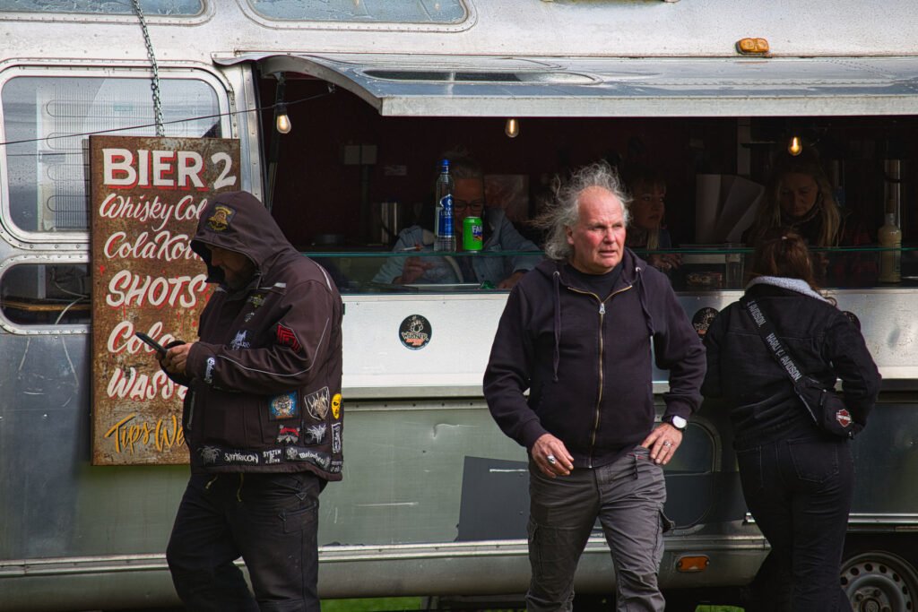 Drei Personen stehen neben einem silbernen Imbisswagen mit einem Schild, auf dem Getränke aufgelistet sind. Ein Mann trägt eine Kapuzenjacke mit Aufnähern und schaut auf sein Handy; ein anderer Mann geht mit einem Getränk spazieren; eine Frau steht vor dem Wagen.