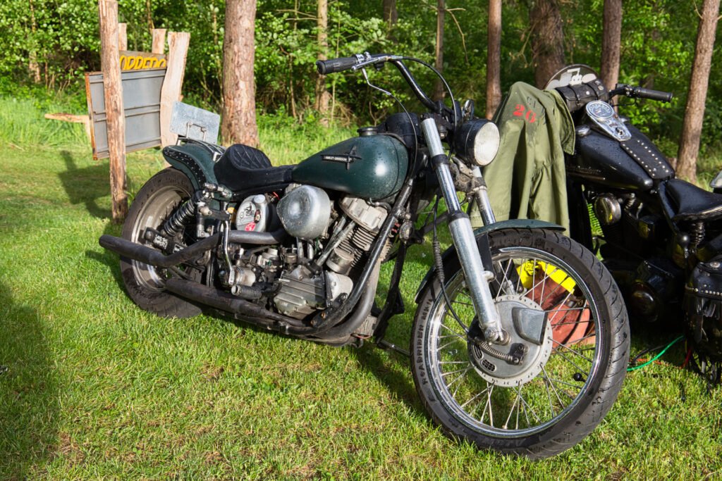 Ein altes grünes Motorrad mit einem abgenutzten schwarzen Sitz ist auf einer Wiese in der Nähe von Bäumen geparkt, im Hintergrund sind ein weiteres Motorrad und einige Outdoor-Geräte zu sehen. Das Sonnenlicht beleuchtet das Motorrad und das nahe gelegene Grün.