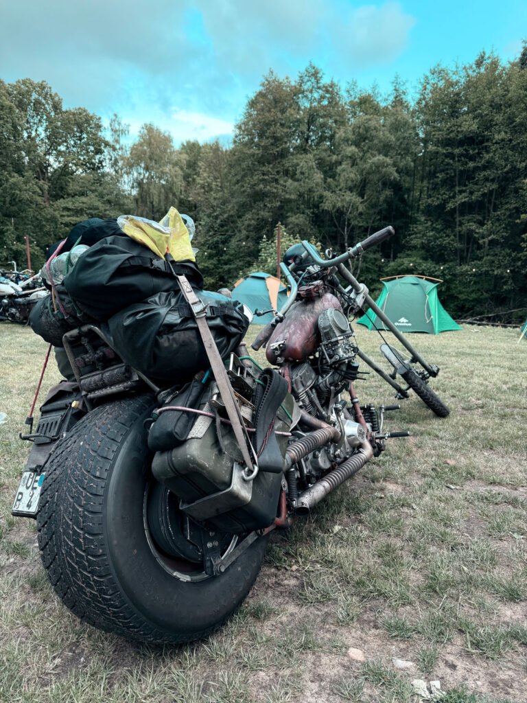 Ein mit Ausrüstung beladenes Motorrad ist auf einer Wiese in der Nähe von grünen Zelten und Bäumen geparkt. Der wolkenverhangene blaue Himmel deutet auf eine Camping- oder Outdoor-Veranstaltung hin.