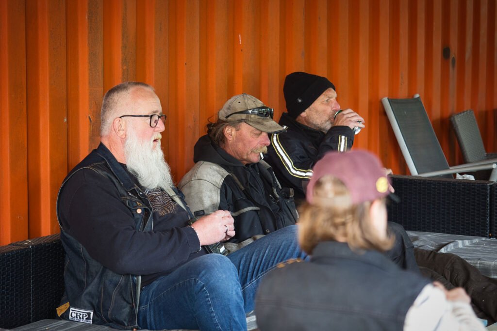 Vier Personen in legerer Outdoor-Kleidung und mit Hüten sitzen auf einer Bank vor einer orangefarbenen Wellblechwand, entspannen sich und trinken aus Bechern. Einige tragen Bärte und Sonnenbrillen, und im Hintergrund sind Gartenstühle zu sehen.