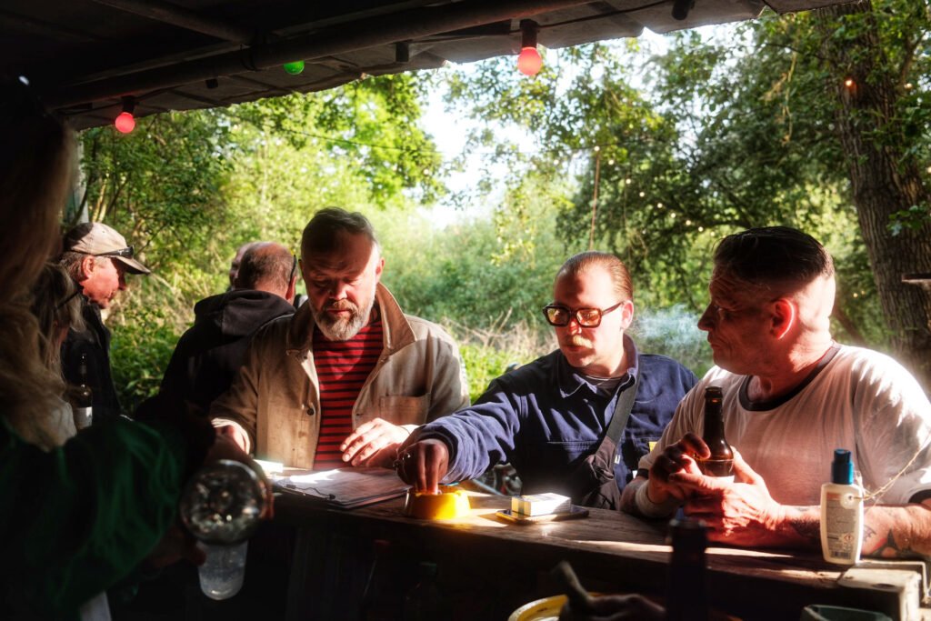 Eine Gruppe von Männern steht an einer hölzernen Bar im Freien unter einem Dach, umgeben von Bäumen und Sonnenlicht. Ein Mann zündet sich eine Zigarette an, ein anderer hält ein Bier in der Hand, und auf dem Tresen liegen Papiere und Getränke. Darüber hängen bunte Lichterketten.