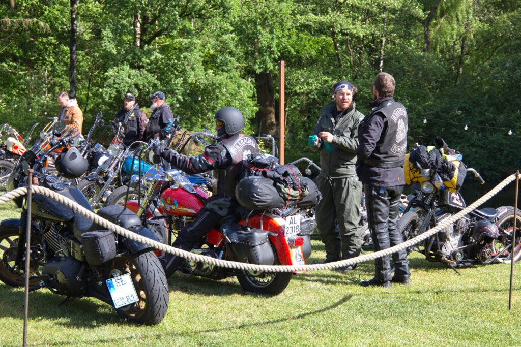 Eine Gruppe von Motorradfahrern in Motorradkleidung steht und unterhält sich in der Nähe von mehreren geparkten Motorrädern auf einer von Bäumen umgebenen Wiese. Einige Motorräder haben Satteltaschen und Reisegepäck.