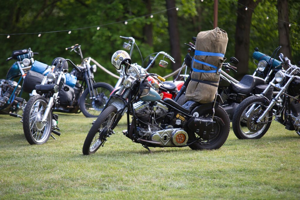 Mehrere geparkte Motorräder auf einer Wiese, wobei ein Motorrad im Vordergrund eine große, braune, aufgerollte Tasche trägt, die senkrecht auf den Rücken geschnallt ist. Im Hintergrund sind Bäume und Lichterketten zu sehen.